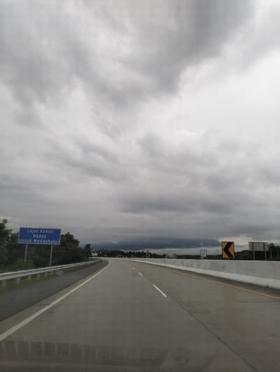 The image depicts a highway scene in Sihomlhok, Indonesia, under a heavily overcast sky, suggesting a cloudy or potentially rainy day. The perspective is from a vehicle traveling on the road, looking forward. The highway itself is paved and appears to be a multi-lane road with clear lane markings. A white guardrail separates the lanes from the shoulder, and a concrete barrier lines the right side of the road.
On the left side of the highway, a blue rectangular sign is visible. The text on the sign reads "Lajur Kanan Hanya Untuk Mendahului", which translates from Indonesian to "Right Lane Only For Overtaking". This indicates that the right lane is designated for passing slower vehicles. Further down the road, on the right side, are several road signs. One is a yellow chevron sign indicating a curve to the left, and another smaller white sign shows "KM 47", indicating the kilometer marker.
The sky is filled with dark, dense clouds, creating a somber and potentially dramatic atmosphere. The horizon in the distance shows a line of trees and possibly some buildings, partially obscured by the low-hanging clouds. The overall impression is one of a journey on a highway in potentially challenging weather conditions, with clear signage providing guidance to drivers. There are no people visible in the image. The time of day is difficult to determine precisely due to the overcast conditions, but it appears to be daytime.
