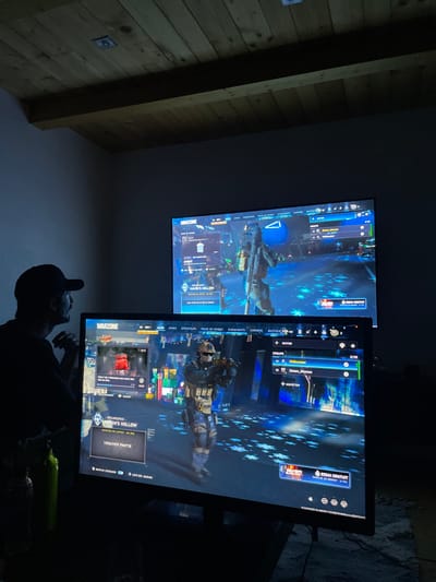 The image shows two screens displaying the game Call of Duty: Warzone. Both screens feature the main menu with a soldier character displayed. The bottom screen shows the game mode "RESURGENCE - HAVEN'S HOLLOW" with an option to "TROUVER PARTIE" (Find Game). The top screen also shows "HAVEN'S HOLLOW" and the same soldier. The menu includes options for "GROUPE" (Group) with a count of 2/32, "COMBAT EVENEMENTS," and "CARRIERE" (Career). In the foreground, a person's silhouette is visible, appearing to be watching the screens.