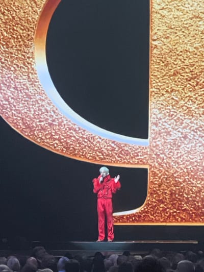 The image displays a performance event within a dark venue in Novoivanovskoe, Russia. A single individual with light-colored, possibly white or light grey, hair is centered on a raised, dark stage. This person wears a prominent red two-piece ensemble, featuring a jacket with ruffled or heavily embellished shoulders and chest, and matching trousers. They are holding a microphone to their mouth with their left hand and have their right hand raised and open in a gesturing motion. Behind the performer is a massive, textured golden backdrop, resembling a large, incomplete letter 'C' or similar curved shape on the left, and a vertical segment on the right. A thin, illuminated line, appearing light blue or white, borders the inner edge of the golden structure. The area below the stage in the foreground reveals the indistinct heads of a seated audience. The scene is illuminated to highlight the performer and the golden backdrop against the otherwise dark environment.