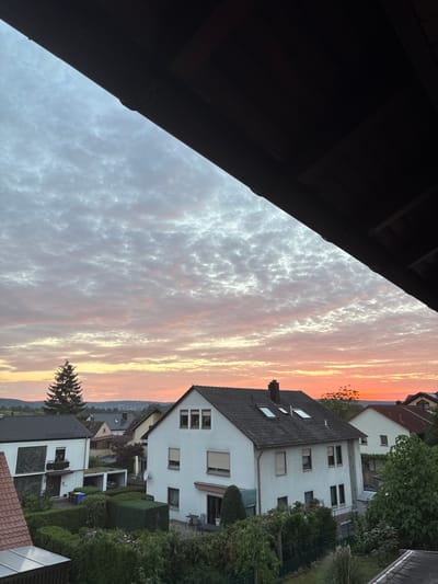 The image captures a view of Thüngersheim, Germany, during a sunset. The sky is filled with a dramatic display of clouds tinged with pink, orange, and white hues. A dark overhang, likely part of a roof or balcony, frames the top of the image, creating a strong diagonal line. Below the sky, a residential area is visible. Several houses with white facades and dark roofs are scattered throughout the landscape. One prominent house in the foreground features multiple windows and a chimney. Green hedges and trees surround the houses, indicating a suburban setting with well-maintained gardens. A tall evergreen tree stands to the left, silhouetted against the colorful sky. The overall scene suggests a peaceful evening in a quiet German town.