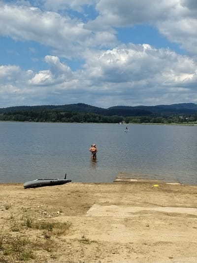 This is a landscape photograph taken at Radslav, Czechia. The scene depicts a large body of water, possibly a lake or reservoir, with a forested hillside in the background. The sky is partly cloudy, with dramatic white and grey clouds against a blue sky.
In the foreground, a sandy shoreline is visible, with a dark-colored watercraft, possibly a kayak or canoe, resting on the sand. Further out on the water, two individuals are standing waist-deep. One individual appears to be embracing or holding another person.
In the middle distance, a lone figure is standing on a paddleboard in the water. The overall impression is a tranquil outdoor recreational setting on a sunny, yet overcast day.