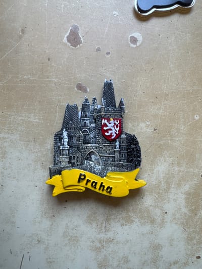 The scene is situated in the city of Neratovice, Czechia. A three-dimensional souvenir magnet is prominently displayed, occupying the lower central portion of the frame. The magnet depicts a detailed rendition of the Charles Bridge and the Old Town Bridge Tower in Prague, featuring grey, stone-like architecture with multiple arched passageways, crenellated walls, and spire-topped towers exhibiting a textured, cross-hatched pattern. Small, sculpted figures resembling statues are visible along the bridge structure. A red shield bearing a white, double-tailed lion rampant with a golden crown, tongue, and claws is affixed to the central tower. Below the architectural representation, a yellow, ribbon-like banner curves upwards, displaying the word "Praha" in raised, black, capitalized lettering. The magnet rests on a flat, light beige or tan surface, which shows numerous fine scratches, scuffs, and several irregularly shaped darker patches, consistent with wear. In the upper right corner of the image, a small portion of a second, dark-colored, cloud-shaped magnet with a metallic border is partially visible and cropped. No individuals are present in the scene.