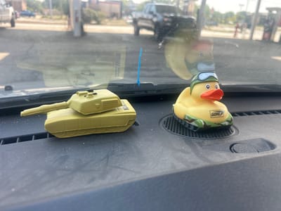 This is a close-up, eye-level shot of two toy military-themed objects on the dashboard of a vehicle, likely a car or truck, parked at a gas station in Willoughby, United States. The dashboard is made of dark, textured plastic and is covered in a fine layer of dust, with some white flecks scattered across it.

On the left is a tan-colored toy tank with a prominent barrel extending to the left. The tank is detailed with visible tracks.

To the right of the tank is a yellow rubber duck wearing a green helmet with blue goggles and an orange beak. The duck is perched on a grey circular grate. The base of the duck toy is camouflaged with green and brown patterns. A small white rectangle with black text, reading "GORE," is visible on the side of the duck's base.

In the background, through the windshield, a gas station is visible. There are gas pumps, a building, and several vehicles, including a dark-colored pickup truck, a white SUV, and a blue sedan. The lighting suggests it is daytime with bright sunlight. A thin, bright blue plastic zip tie stands upright on the dashboard in front of the windshield. The reflection of the outside environment is visible on the windshield, including what appears to be the silhouette of the tank and possibly the duck.