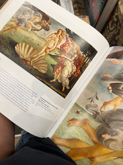 An open art history book, held on a person's lap, displays reproductions of classical artworks. The left page features Sandro Botticelli's "The Birth of Venus," depicting Venus emerging from a scallop shell, accompanied by Zephyrus and Hora. Text below identifies the painting as "The Birth of Venus, 1484-1485" by Sandro Botticelli, noting its dimensions, medium (tempera on canvas), and current location at the Uffizi Gallery. The right page shows a partial, close-up reproduction of what appears to be another Botticelli painting, likely "Primavera," with visible bare feet, hands, flowing drapery, and scattered pink flowers against a light green and blue background. The person holding the book wears dark blue denim shorts and a black t-shirt. The background, out of focus, reveals bookshelves or display racks filled with other books, indicating a retail environment such as a bookstore. No specific visual identifiers are present in the image to pinpoint a precise location within the city of El Paso, United States.