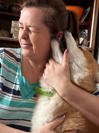 A woman and a dog are closely interacting in an indoor setting. The woman has her eyes closed and her face slightly contorted, appearing to either be squeezing her eyes shut or smiling. She's wearing a striped shirt of turquoise, white, and navy blue, with a solid turquoise top underneath. The dog is a light brown color and has a green collar. The dog's face is close to the woman's head, and she has her hand around its neck, possibly hugging it. The environment is dimly lit, and details behind the figures are not clearly discernible, but it includes what appears to be shelving or boxes. There is no information related to Заболотово, Russia, in the image.