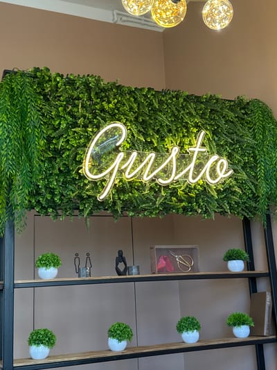 The image shows a neon sign that spells "Gusto" in cursive, glowing white, mounted on a wall covered in artificial greenery. The sign is positioned above two shelves of a dark metal shelving unit. The top shelf contains two small potted plants, a set of two stylized human figures, a decorative object of a person in a thoughtful pose, and a small wooden box with fabric and scissors inside. The second shelf holds one small potted plant. The third shelf has two small potted plants and a book. The background wall is a neutral beige color. The scene is illuminated by warm, ambient light, possibly from overhead fixtures visible at the top of the image. The location of Патрушева, Russia is not discernible from the image content.