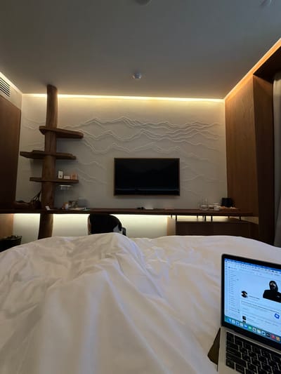 The image shows an interior scene. A bed with white linens takes up a large portion of the right side of the frame. A laptop rests on the bed, displaying a webpage in Russian. To the left of the bed, a wall features decorative white lines and a flat-screen television mounted. Below the TV is a wooden structure, possibly a shelf or decorative element. The room is illuminated by recessed lighting along the wall. There is nothing to indicate this image was taken in the city of Озёрное, Russia.