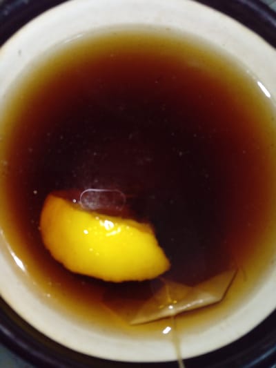 The image shows a close-up overhead view of a mug of dark tea. A slice of bright yellow lemon floats on the surface of the tea, positioned to the left of the center of the mug. Partially submerged in the tea near the lemon slice is a tea bag, its paper tag folded and visible just below the liquid's surface. The mug itself is light-colored with a beige rim, and it sits within a darker outer container, possibly a coaster or another mug. The lighting casts reflections on the surface of the tea, creating bright, circular highlights. The scene is static and appears to be indoors, with no discernible elements that would place it specifically in Kitskany, Moldova.