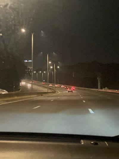The image captures a nighttime view from an elevated highway or bridge, likely in Hod HaSharon, Israel. The scene is dark, illuminated primarily by artificial lights. The road surface appears wet and reflective, suggesting recent rain or ongoing light precipitation. White dashed lane markings are visible on the multi-lane road, which stretches into the distance.
On the immediate right, a dark, vertical structure, possibly a railing or bridge support, frames a portion of the image. Further along the highway, the red tail lights of several vehicles are visible, indicating traffic moving in the distance. To the left of the road, numerous streetlights line an area below the highway, casting reflections and illuminating a landscape that includes distant building lights, characteristic of an urban or semi-urban environment. The sky above is featureless and dark. No people are visible, and the scene conveys a calm, functional atmosphere typical of night travel. There is no visible text in the image.