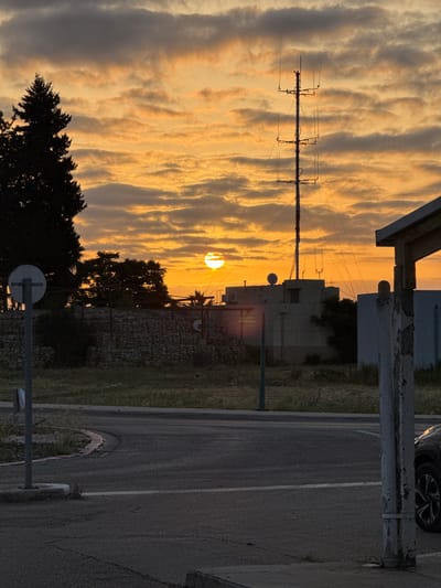 The image depicts a sunset in Mazkeret Batya, Israel. The sun is setting behind a large, multi-sectioned antenna pole and a concrete building with a wall in front of it. The building has a small satellite dish on top. In the foreground there is a paved street with a crosswalk and a stop sign. There is a tree in the lower left corner. The sky is orange and yellow, with some clouds. A car is visible in the lower right corner of the image.