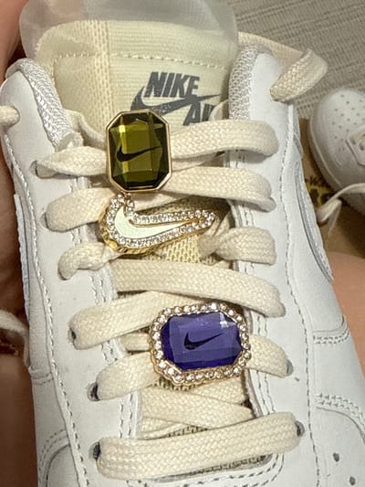 This image depicts a close-up view of a white Nike Air Force 1 shoe. The laces are also white and adorned with three decorative charms. The charm on the left is a dark green octagon with a gold border and a Nike swoosh design. The charm in the center is a curved Nike swoosh shape with a gold border and inlaid with small stones or rhinestones. The charm on the right is a blue octagon with a gold border featuring small stones or rhinestones around its edge, also containing a Nike swoosh design. The surrounding environment is out of focus, suggesting a domestic setting. The location of Hainburg an der Donau, Austria, is not discernible from the image.