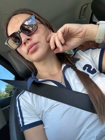 A young woman is taking a selfie in a car. She is wearing sunglasses, a white t-shirt with blue trim and a number "2" on it, and a watch on her left wrist. Her brown hair is tied in a ponytail. A seatbelt is across her chest. The scene appears to be outdoors during the daytime, as suggested by the sunlight and the reflection of buildings in her sunglasses. The woman's expression is neutral, and she is looking directly at the camera. The setting seems to be a car interior, with a black leather seat visible. The overall mood is casual and relaxed.