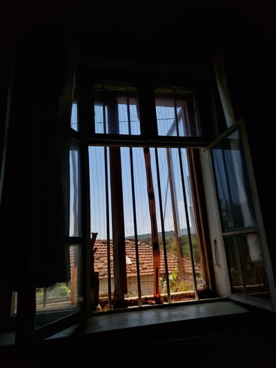 The image depicts an interior view looking out of a window. The window frame is old and appears weathered, with some damage visible on the right-hand side. The window is partially open, and through the bars, a rural landscape can be seen. The landscape includes a building with a tiled roof, trees, and a clear blue sky. The interior of the room is dark, with a heavy curtain visible on the left side, obscuring a large portion of the view. The overall impression is one of abandonment or neglect. The image was captured in Raduvene, Bulgaria, a town known for its rural setting and traditional architecture.