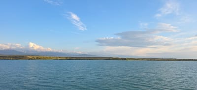 This is a scenic, wide-angle, landscape photograph taken from the shore of a large body of water, likely Issyk-Kul Lake, near Pristan'-Przheval'sk, Kyrgyzstan. The foreground is dominated by the rippling, turquoise surface of the lake, reflecting the bright blue sky. The horizon line divides the image, with the distant shore featuring a strip of green land dotted with sparse vegetation and some scattered buildings. Beyond the land, a majestic mountain range stretches across the background, its peaks capped with snow and illuminated by the soft light of the setting sun. Wispy white clouds drift across the expansive sky, adding depth and texture to the scene. The overall impression is one of tranquility and natural beauty. There are no people or animals visible in the image.