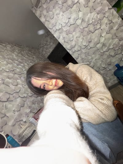 The image shows a young woman with long brown hair sitting indoors, her head tilted down and resting on her arm. She is wearing a cream-colored knitted sweater and blue jeans. The background is decorated with floral wallpaper. On a surface next to her, several items are visible, including a blue bottle, a makeup bag, a comb, and what appear to be some small containers or pills. To the right of the woman, a large blue water bottle is partially visible. The lighting suggests an indoor setting, possibly a home. No specific landmarks of Sakmary, Russia, are discernible in the image.