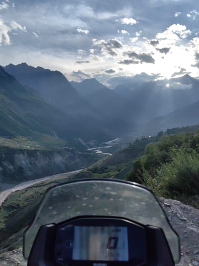 The image shows a mountainous landscape with a river flowing through a valley. The foreground is dominated by a motorcycle windshield, partially obscuring the view of the landscape. The windshield is clear with some water spots. Beneath the windshield, a digital display shows a "0" and other unreadable indicators.

The landscape is characterized by steep, verdant slopes on either side of the valley. The mountains in the background are tall and rugged, shrouded in a soft haze. The sky is partly cloudy, with the sun breaking through the clouds, casting dramatic rays of light that illuminate parts of the valley. A river snakes through the valley floor, appearing as a pale ribbon. Small settlements with houses are visible on the slopes, nestled amongst the greenery. The terrain in the foreground is rocky and uneven.

The overall impression is one of a scenic, remote location, possibly during late afternoon given the quality of light. The perspective suggests the photo was taken from a moving vehicle, likely the motorcycle in the foreground. The presence of the motorcycle and its instruments indicates a journey or exploration.