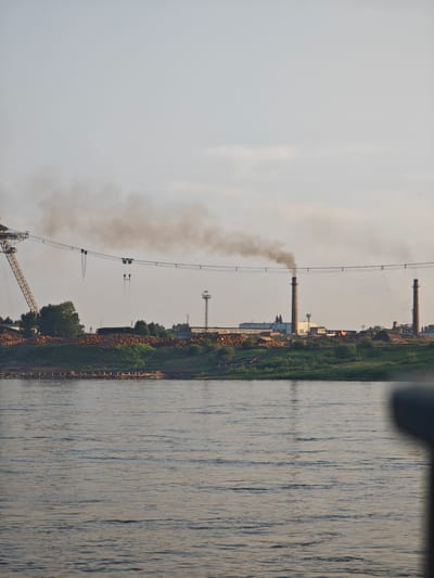 The image captures a scene on the Yenisei River in Lesosibirsk, Russia. The river occupies the foreground, its surface rippled by a gentle current. On the far bank, a sprawling industrial complex is visible. Prominent structures include several tall brick chimneys, one of which is emitting a thick plume of dark smoke into the hazy sky. In the middle ground, a large quantity of logs is stacked along the riverbank, suggesting a lumber or wood processing facility. To the left, a large yellow industrial crane stands, its boom extended over the area. The background is filled with the muted tones of a hazy sky and distant trees. There are no people visible in the image.