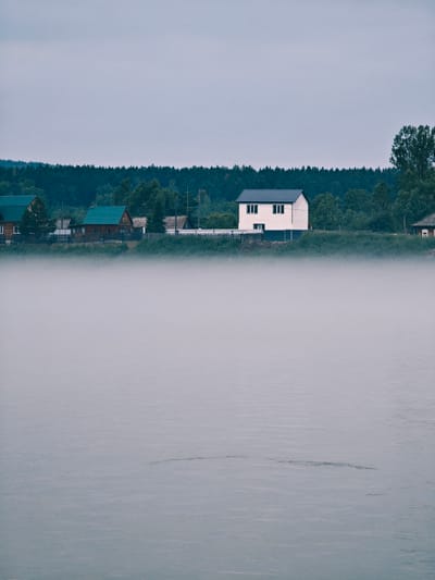 This image depicts a serene scene in the city of Atamanovo, Russia. In the foreground, a body of water is shrouded in a dense layer of fog, its surface disturbed by subtle ripples. Beyond the fog, the opposite bank of the water features a line of houses nestled against a backdrop of dense green forest. Several houses with dark roofs are visible, including a prominent two-story white house with dark-trimmed windows. A wooden fence runs along the bank in front of some of the houses. The sky above is overcast and a pale gray, contributing to the subdued and atmospheric mood of the scene. The overall impression is one of quiet and natural beauty.