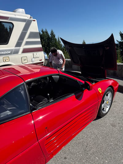 The image captures a bright, sunny outdoor scene featuring a red Ferrari sports car and a recreational vehicle (RV) parked on asphalt pavement. The Ferrari, likely a classic model like a 348, is positioned prominently in the foreground and midground. It is a striking red color with silver five-spoke wheels, each featuring a yellow Ferrari emblem on the hub cap. A smaller yellow Ferrari badge is also visible on the front fender. Distinctive horizontal strakes adorn the side bodywork. The car's front hood is open, and a man is leaning over it, peering into the compartment.
The man is wearing a white polo shirt with green and red striped trim on the collar and sleeves, and sunglasses, appearing focused on inspecting or attending to the car. To the left and slightly behind the Ferrari is a large, light-colored RV with prominent gray and maroon graphic stripes along its side. The brand name "CHINOOK" is partially visible on the side of the RV.
The setting is an open area under a clear, bright blue sky, suggesting excellent weather, likely during the day. Green trees are visible in the background, hinting at a natural or park-like environment in West Vancouver, Canada. Visible text includes "CONTINENTAL" on the Ferrari's tire and "G2E255LUR" on its passenger side window. The interior of the Ferrari shows a black steering wheel, seats, and a metallic gear shifter.