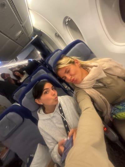 Two women are seated in adjacent airplane seats, posing for a selfie. The woman on the left has blonde hair and is wearing a tan jacket and scarf. The woman on the right has dark hair and is wearing a gray hooded sweatshirt. Both women are making kissing faces. The plane cabin is visible in the background, with other passengers and rows of seats. The image is slightly tilted.
