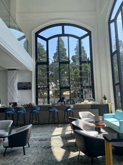 This image captures the interior of a brightly lit bar or lounge area, specified to be in Kayuambon, Indonesia. The space features high white ceilings and walls, with natural light entering through multiple tall, arched, black-framed windows spanning the back wall. Outside the windows, numerous green coniferous trees are visible under a clear blue sky.

In the center of the frame, a dark-toned bar counter runs horizontally, equipped with several dark blue velvet bar stools with black legs and gold accents. Behind the bar, a gold-colored, arched shelving unit filled with various glasses and bottles is positioned in front of the central window. A male individual in a dark shirt is partially visible behind the bar on the left, appearing to be working. On the bar counter, various bottles of spirits, bar tools, and small potted plants are arranged. To the left of the bar, a large fluted column with a marble-like finish supports the structure, and above it, a flat-screen television displaying an image of a vehicle is mounted on the wall.

The foreground and mid-ground feature a lounge seating arrangement on a large rug with a mottled grey and white pattern. Several armchairs are present: a light grey upholstered armchair, multiple grey armchairs with black exterior frames, and a light blue upholstered sofa to the right. A round, dark wooden coffee table is placed among the grey armchairs, and a rectangular black table with a gold base is positioned near the blue sofa. Strong sunlight casts distinct shadows of the furniture across the rug from the right side of the frame. Through a window on the far right, a partial blue sign with white text, including the word "PARKING," is discernible, indicating an outdoor area adjacent to the building. A small potted plant sits on the bar counter to the right.