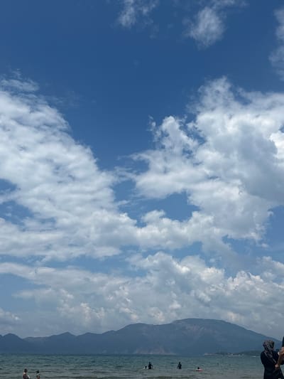 The image depicts a seascape in Ortaca, Turkey. To the left, the sky is a deep blue, gradually transitioning to a mix of white cumulus clouds interspersed with patches of lighter blue. The sea is visible on the right side, reflecting the sunlight and showing small waves. Along the shoreline, there are a few people in the water, appearing to be swimming or engaging in water activities. In the background, a mountain range rises steeply. The overall lighting indicates a bright, sunny day.