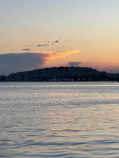 This image captures a coastal city at dusk, likely within the Municipal Unit of Dafni, Greece, given the hilly terrain densely populated with buildings and its proximity to the sea. The foreground is dominated by the rippling surface of the water, reflecting the soft hues of the sunset. Beyond the water, a hillside city is silhouetted against the sky, its buildings clustered together, creating a textured, dark mass. Several yachts and boats are moored or sailing in the harbor area along the coastline. The sky above is a gradient of soft blues and oranges, with scattered clouds, some of which are illuminated by the setting sun, adding a touch of dramatic color. The overall atmosphere is one of peaceful transition from day to night.