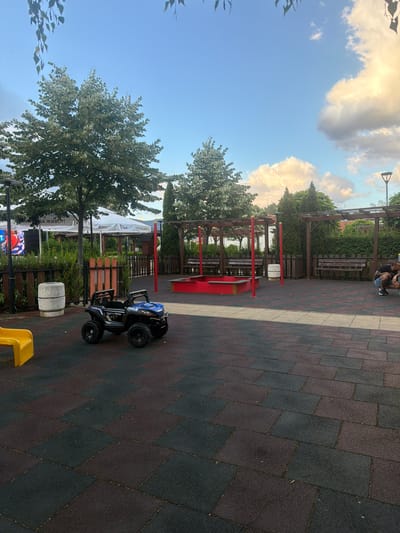 An outdoor scene on a playground in Chelopech, Bulgaria, captured during the day. The ground is paved with interlocking rubber tiles in dark red and black. In the foreground, a blue toy electric car is parked. To the left, a portion of a yellow slide is visible. Behind the car, a circular concrete planter with a rough texture stands. Further back, a red sandbox with wooden borders is situated under a wooden pergola structure. Benches made of wood are placed along the back of the playground. The background features lush green trees and bushes, and a large white tent with writing on it. The sky above is blue with scattered white and golden clouds. A street lamp is visible on the right side of the image. A man is crouched down on the far right, seemingly looking at something on the ground.