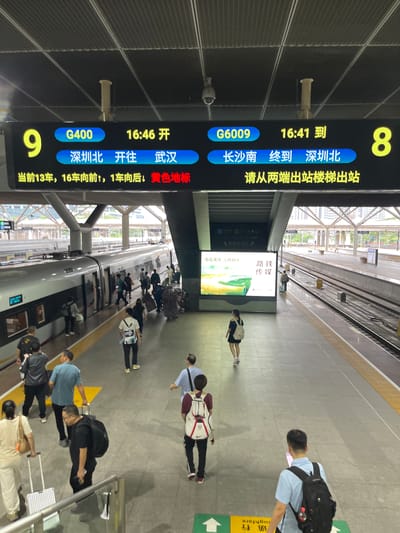 This image captures a bustling train station platform in China, likely during the daytime given the lighting. The primary focus is on a digital display board showing train information and the people waiting on the platform.
The digital display board prominently features train numbers like "G400" and "G6009," departure and arrival times ("16:46 开", "16:41 到"), and destinations ("深圳北 开往武汉", "长沙南 终到 深圳北"). It also includes directional information for passengers, such as "当前13车,16车向前,1车向后,黄色地标" and "请从两端出站楼梯出站". A train is visible on the tracks, suggesting an active station environment.
Several people are scattered across the platform, some with luggage, indicating travel. Their postures suggest waiting, moving, or possibly orienting themselves with the information provided. One person is looking at their phone, while others are walking or standing. The presence of luggage, backpacks, and shopping bags highlights the travel context.
A large advertisement panel with Chinese text, including "路铁 传媒" and "旅途漫漫之明相伴", is also visible, adding a layer of commercial or promotional content to the scene. The setting appears to be a modern, covered train station with clean gray flooring and visible ceiling structures. The overall atmosphere is one of transit and everyday activity at a busy transportation hub.