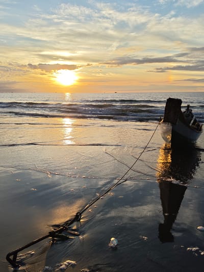 The image depicts a tranquil beach scene in Uleekareung, Indonesia, during sunset. The sky is painted with warm hues of orange and yellow, with scattered clouds adding texture to the canvas. The sun, partially obscured by clouds, casts a golden reflection on the calm sea, where gentle waves lap at the shore. In the foreground, a small boat rests on the wet sand, its anchor rope taut, connecting it to an anchor embedded in the sand. The wet sand reflects the warm light of the setting sun, creating a shimmering effect. The scene conveys a sense of peace and serenity, characteristic of a sunset on a tropical beach.