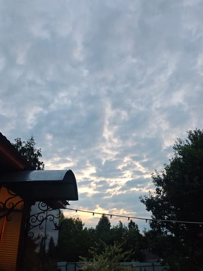 A medium shot captures a twilight sky over a wooded area and a house in Luzhniki SNT, Russia. The sky is filled with a pattern of scattered clouds, with hints of the setting sun peeking through in the distance, casting a soft glow. In the foreground, a portion of a house is visible on the left, featuring a dark awning with decorative metalwork and a string of lights stretched across the frame towards the right. Dense trees form a dark silhouette against the sky, with some foliage encroaching on the edges of the frame. The overall mood is serene and peaceful, indicative of the end of the day.