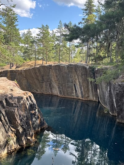 The image shows a quarry in Säter, Sweden, with steep, rocky walls surrounding a dark, reflective body of water. Pine trees line the top of the quarry, with their reflections visible in the water. The sky above is blue with scattered white clouds. The rock faces are textured and show layers of different colors, primarily brown and gray, with some white mineral deposits. The water is a deep blue-green and appears still, mirroring the surrounding trees and sky. In the foreground, to the left, a prominent rock formation juts out, also showing textured, layered surfaces. There are no people or animals visible in the image. The overall impression is one of a natural, somewhat rugged landscape.