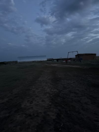 The image depicts a rural landscape under a cloudy, twilight sky in Бөрлі ауылдық округі, Kazakhstan. In the mid-ground, a large, elongated, metallic structure, possibly a greenhouse or storage facility, is visible on the left. To the right of this structure, a small, rustic building with a thatched or corrugated roof and a swing set with a red and white sign in front of it are present. The foreground is a dirt road or track, leading towards the structures. The overall impression is one of a remote, agricultural or semi-agricultural setting.