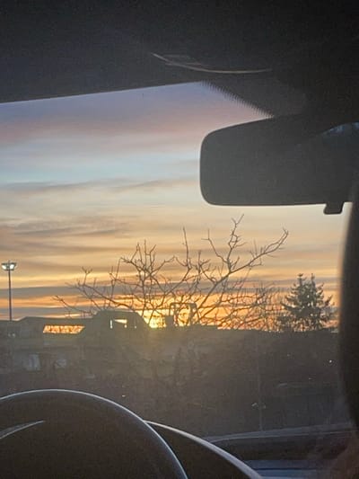The image depicts a golden sunrise or sunset viewed through a car window. Silhouetted branches of a tree reach upward against the sky's vibrant hues. A streetlight stands below. Interior car elements, including the rearview mirror and the steering wheel, frame the outdoor scene, creating a sense of being inside a vehicle.