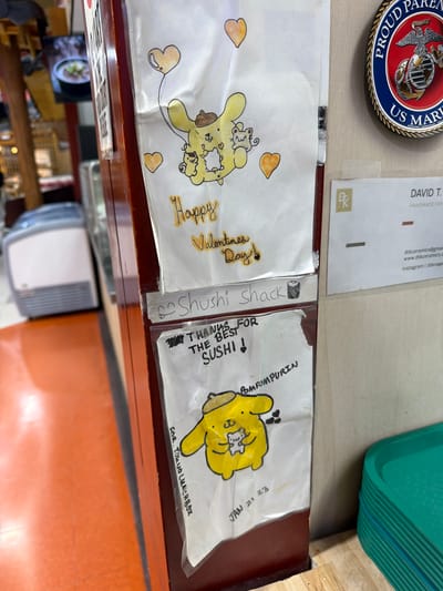 The image shows a close-up shot of a countertop in a place called "Shushi Shack," likely a sushi restaurant or store. The countertop appears to be made of wood, with a reddish-brown color. On the countertop are two sheets of paper with drawings on them. One paper has the words "Happy Valentines Day!" and a cartoon drawing of a yellow character surrounded by hearts. The other paper says "THANKS FOR THE BEST SUSHI!" with a drawing of a yellow cartoon character labeled "POM FOMPURIN." It also has the text "FOR. TOKUO LUNCHBOX" and a handwritten date "JAN 21 23." Additionally, there's a small slip of paper with a drawing of a sushi roll. Above the papers are several other items. A military coin with the words "US MARINE PARENTS" is on the left side, and a business card that says "DAVID T. HANDMADE CERAMICS" is on the right. A green container is visible in the upper right corner. The lower part of the image shows a shelf with various items. The photo was taken in Niles, United States of America, based on the OCR.