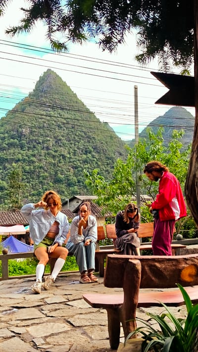 In the Sà Phìn Commune, Vietnam, a group of four individuals are situated outdoors, amidst a hilly landscape. Three women are seated on a wooden bench, with a man standing to their right. One woman, positioned on the far left, wears a light blue jacket, green shorts, and white knee-high socks. The woman beside her is dressed in a light blue shirt and jeans. The third woman is wearing dark clothes and looking at her phone. The man is attired in a red jacket and maroon pants. The background features lush green mountains and residential structures with roofs. Power lines are visible against the sky. The foreground includes a paved area and a park bench made of wood, around which greenery and plants grow.