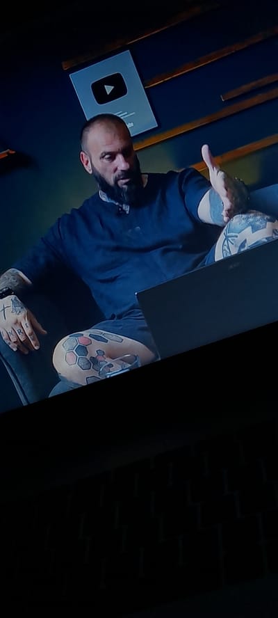 In the city of Zlataritsa, Bulgaria, a man with a shaved head and beard, adorned with numerous tattoos on his arms and legs, is seated. He is wearing a dark blue t-shirt and appears to be engaged in a conversation, gesturing with his right hand. In front of him rests a dark-colored laptop. Behind him, a silver YouTube play button award is mounted on a wall with dark blue paneling and wooden slats. The lower portion of the image is dark and indistinct, showing a patterned surface.