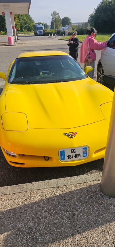 A yellow Chevrolet Corvette C5, bearing French license plate EG-187-AM, with the Brittany region and department 35 (Ille-et-Vilaine) identifier, is parked on a speckled concrete surface with a concrete curb. The front bumper shows some minor dirt and an exposed tow hook receiver. A black and white checkered Corvette emblem is affixed to the front.

To the left of the Corvette, a person with dark hair and glasses, dressed in a black top, holds a smartphone horizontally, appearing to photograph or video. To the right, a second person with dark hair tied back, wearing a pink fluffy top and light-colored pants with a multi-colored shoulder bag, leans towards the open passenger window of a partially visible white vehicle.

In the background, a gas station canopy with white pillars and red pump numbers (1 and 2) is visible on the left. Pink trash receptacles are positioned near the pumps. Beyond the gas station, a blue Scania semi-truck is visible on a paved area. The mid-ground features a road, traffic cones, and dense green foliage. Further in the background, more trees and a distant light-colored building are present under a clear sky. A metal pole casts a shadow on the right foreground. The scene is illuminated by natural daylight, casting distinct shadows. This location is identified as being within the city of Bain-de-Bretagne, France, based on provided contextual information.