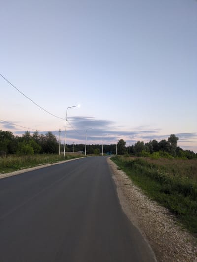 The image shows a paved road curving to the right, leading into a landscape of trees and vegetation under a pale blue sky with wispy clouds. There are several lampposts lining the right side of the road, with one in the foreground actively illuminated. The road surface appears to be asphalt, with a gravel shoulder on the right. The left side of the road is bordered by a grassy area with scattered trees and a wooden fence. In the distance, there is a dense line of trees forming a forest. The overall scene suggests a rural setting, likely in the evening due to the illuminated lamppost and the twilight sky. This image is identified as being in the village of Posyolok Molochnoy Fermy, Russia.