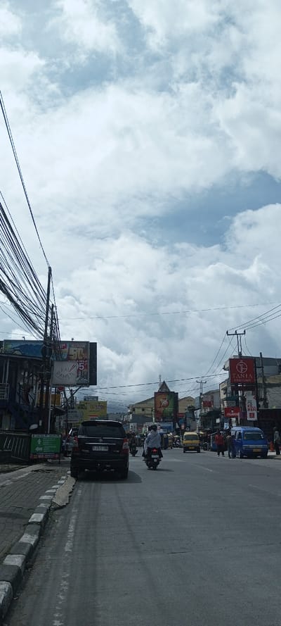 An urban street scene under a cloudy sky features a mix of vehicles including cars, motorcycles, and vans. Utility wires tangle overhead. Buildings line the sides of the street, adorned with billboards and commercial signs. Pedestrians can be seen near the sidewalk. The road stretches into the distance, with hints of mountains visible in the background.