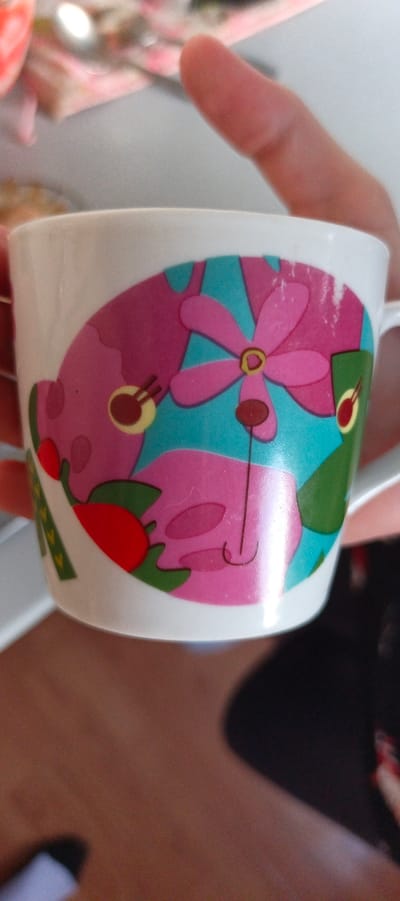 The image shows a close-up of a white ceramic mug with a colorful, abstract design. The design features large, stylized pink flowers with yellow centers against a bright turquoise background. There are also abstract shapes in pink and green, and a small red oval shape. A thin black line, possibly representing a stem or a drawn element, curves across the lower part of the design. The mug is held by a person's hand, with fingers visible on the left and a thumb on the right. The background is blurred, but appears to be an indoor setting with a wooden surface below and a lighter surface above, possibly a table or countertop. The lighting suggests it is daytime. The overall impression is of a playful and decorative object. There is no visible text.