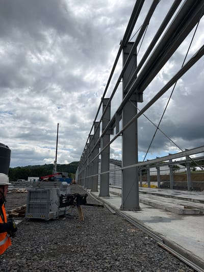 The image depicts a construction site with a focus on the steel framework of a building. The structure consists of large grey metal beams and columns, some of which are angled and supported by diagonal braces. The ground is covered with gravel, and concrete slabs are laid out in a long, linear fashion. In the foreground, a construction worker wearing a white hard hat and an orange safety vest is visible on the left, facing away from the camera. To the right of the worker, there is a grey metal cage-like structure with a mechanical arm attached. In the background, a tall crane with a black telescopic boom is positioned, alongside a red Manitou telehandler. Further back, there are more industrial buildings and a landscape of rolling green hills under a cloudy sky. The location is identified as Jodłownik, Poland.