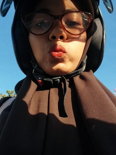 This is a selfie taken by a person wearing a helmet and glasses. The subject is a young person, likely female, with visible makeup on their lips. They are wearing a brown hijab and a brown outer garment, possibly a jacket or sweater. The helmet is black with a textured surface and a chin strap that is fastened with a black and red clip.

The photo appears to be taken outdoors on a clear, sunny day. The sky is a vibrant blue, and the lighting suggests it is daytime, likely late morning or early afternoon due to the angle of the sun and the shadows cast. There are some trees visible in the lower right portion of the frame, suggesting a somewhat natural or park-like setting.

The perspective is from a low angle, looking up at the subject, which is common in selfies. The subject's face is the primary focus, with their lips slightly parted and their eyes visible through their glasses. The overall mood seems casual and possibly adventurous, given the helmet. There is no visible text in the image. The location context is Macege, Indonesia.