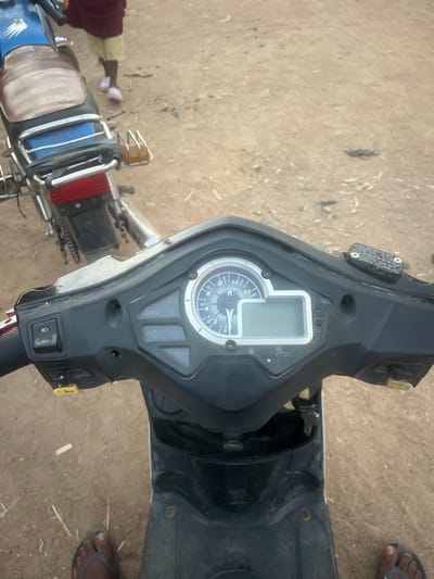An overhead shot showcases the handlebar and dashboard of a scooter. The instrument panel features a digital display and an analog speedometer. To the right, the scooter's body is visible, along with a glimpse of a person's foot wearing flip-flops. On the left, parts of another motorcycle are visible, including its seat, rear light, and luggage rack. The ground serves as the backdrop.