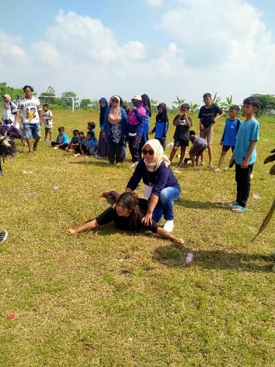 An outdoor scene in Celep, Indonesia, depicts a group of individuals gathered on a sunny, grassy field under a bright blue sky with scattered white clouds. In the foreground, a young woman with dark hair, wearing a dark long-sleeved shirt, lies prone on the grass with her arms outstretched to the sides. An adult woman, wearing a light-colored hijab, sunglasses, a dark long-sleeved top, blue jeans, and white sneakers, kneels over the prone woman, placing her hands on her back/shoulders, appearing to assist or guide her. In the mid-ground and background, a group of approximately 15-20 adults and children observe the activity. This group includes individuals in various casual attire; some women wear hijabs, while others wear t-shirts, shorts, and jeans. Some children are seated on the grass, while others stand watching. Notable figures in the background include an adult male in a white "GARFIELD 22" t-shirt and blue shorts walking to the left, and a young boy in a light blue t-shirt standing on the right. A white soccer goalpost is visible in the far-left background, beyond which are trees and foliage. Small pieces of litter are scattered across the grass.