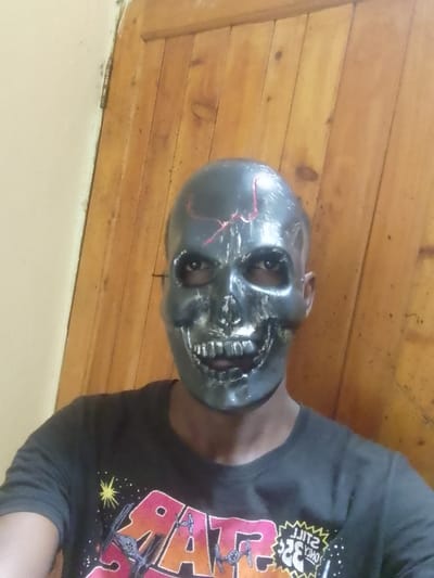 A single individual is depicted indoors in the city of Ziwa La Ng'ombe ward, Kenya. The subject wears a dark grey, skull-shaped mask with visible eyeholes revealing human eyes. The mask features a simulated nasal cavity and exposed teeth in the mouth area. Red, cracked-line patterns are present on the mask's forehead and upper facial regions. The individual is wearing a black t-shirt with a graphic design featuring the "STAR WARS" logo, various starships, and the mirrored text "STILL HERE." The background consists of a light beige wall on the left and a vertical-plank wooden door or paneling on the right, with a visible metallic hinge connecting it to the wall. The subject is oriented directly towards the camera.