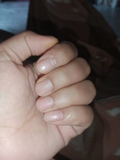 The image displays a close-up of a person's hand, with the fingers curled inward. The skin appears to be of a light to medium complexion. The fingernails are short and unpolished, showing a natural, light pink hue. The lighting suggests an indoor setting, possibly with artificial light casting subtle shadows. The background is out of focus and appears to be a textured fabric in muted tones of brown and beige. There is no indication of any specific location within the city of Cotseutui, Indonesia, as the image focuses solely on the hand and its immediate surroundings. The scene is static, with no discernible action or interaction taking place.