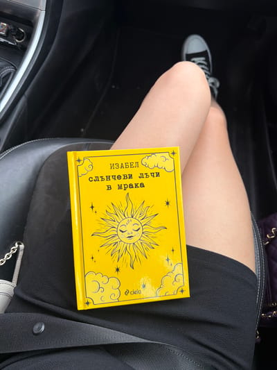 A person is seated inside a vehicle, holding a bright yellow book with a sun illustration and text in Bulgarian. The book's title appears to be "Слънчеви лъчи в мрака" (Slanchevi lachyi v mraka), translating to "Sunbeams in the Dark." The individual is wearing a black dress and black Converse sneakers. The setting is the interior of a car, with visible elements of the dashboard and seat upholstery. The car is parked in Aleksandrovo, Bulgaria.