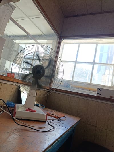 The image shows an indoor scene, likely a room with a window. A white oscillating fan with a metallic grille is positioned on a wooden table in the foreground. The fan is in operation, as indicated by the blurred blades. The table is cluttered with various items, including electrical cords, a blue plastic bag, and a small red object that may be a button or tag. The room features tiled walls and a wooden ceiling with visible beams and conduits. Through the window, a bright outdoor scene is visible, with a grid-like structure and what appears to be a building or industrial area in the distance. The overall impression is of a functional, somewhat utilitarian space. Without further information or context, it is not possible to definitively place this scene in Izberbash, Russia, or any other specific location.