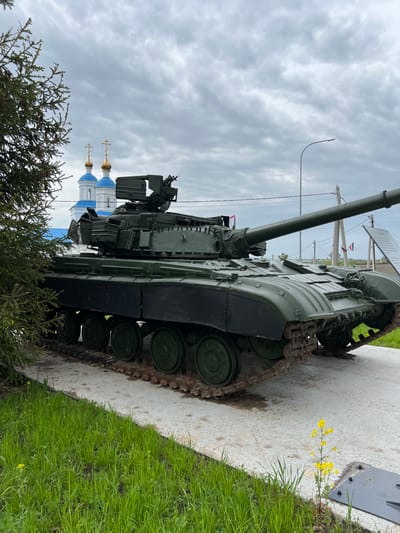 A tank is displayed outdoors in Введенская Слобода, Russia. A grassy area and a concrete sidewalk are visible to the right. An evergreen tree is to the left. In the background, there are two buildings with gold crosses, a street lamp, and clouds in the sky.